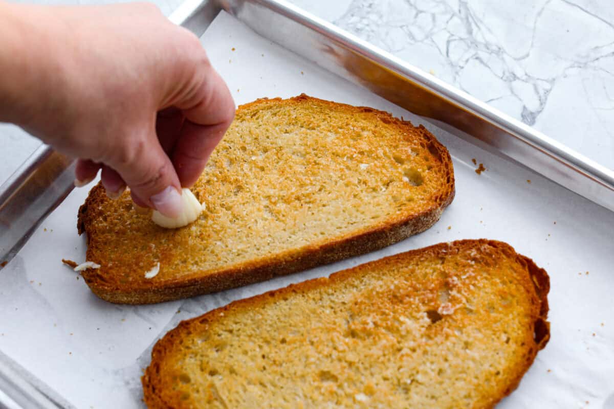 Vibrantย Tomato Toastย has sweet, juicy tomatoes, garlic, creamy ricotta, and fresh basil layered on a crusty slice of toasted bread. Itโs delicious and savory with hints of sweetness. Serve it for breakfast, brunch, as an appetizer, or light lunch for the perfect way to use up summer produce. Overhead shot of someone rubbing garlic clove on top of toasted bread.