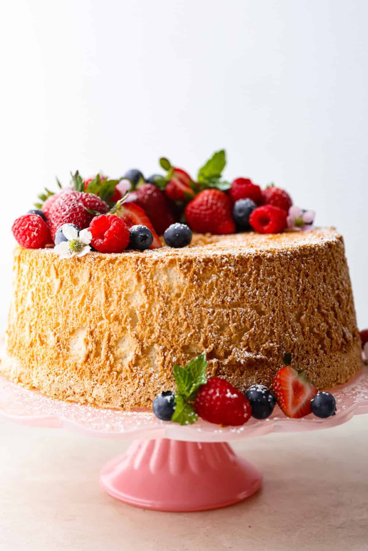 This light & fluffy Angel Food Cake is like a slice of heaven on a plate! It's delicious all on its own but absolutely divine topped with sweetened whipped cream and fresh berries or lemon curd! Side shot of whole angel food cake with berries and blossoms dusted with powdered sugar.
