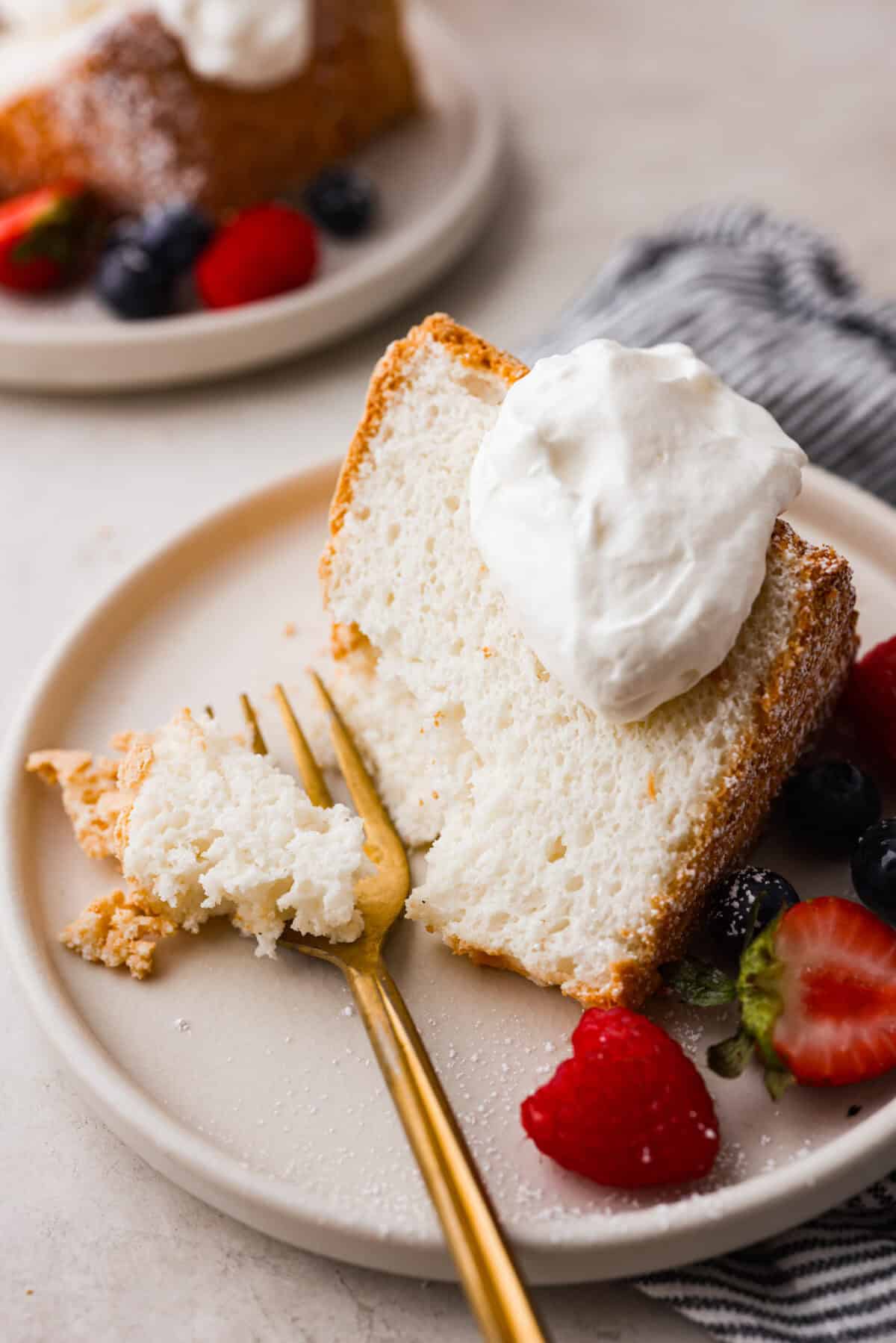 This light & fluffy Angel Food Cake is like a slice of heaven on a plate! It's delicious all on its own but absolutely divine topped with sweetened whipped cream and fresh berries or lemon curd! Angle shot of plated angel food cake with dollop of whipped cream on top and berries on the side, next to a gold fork.