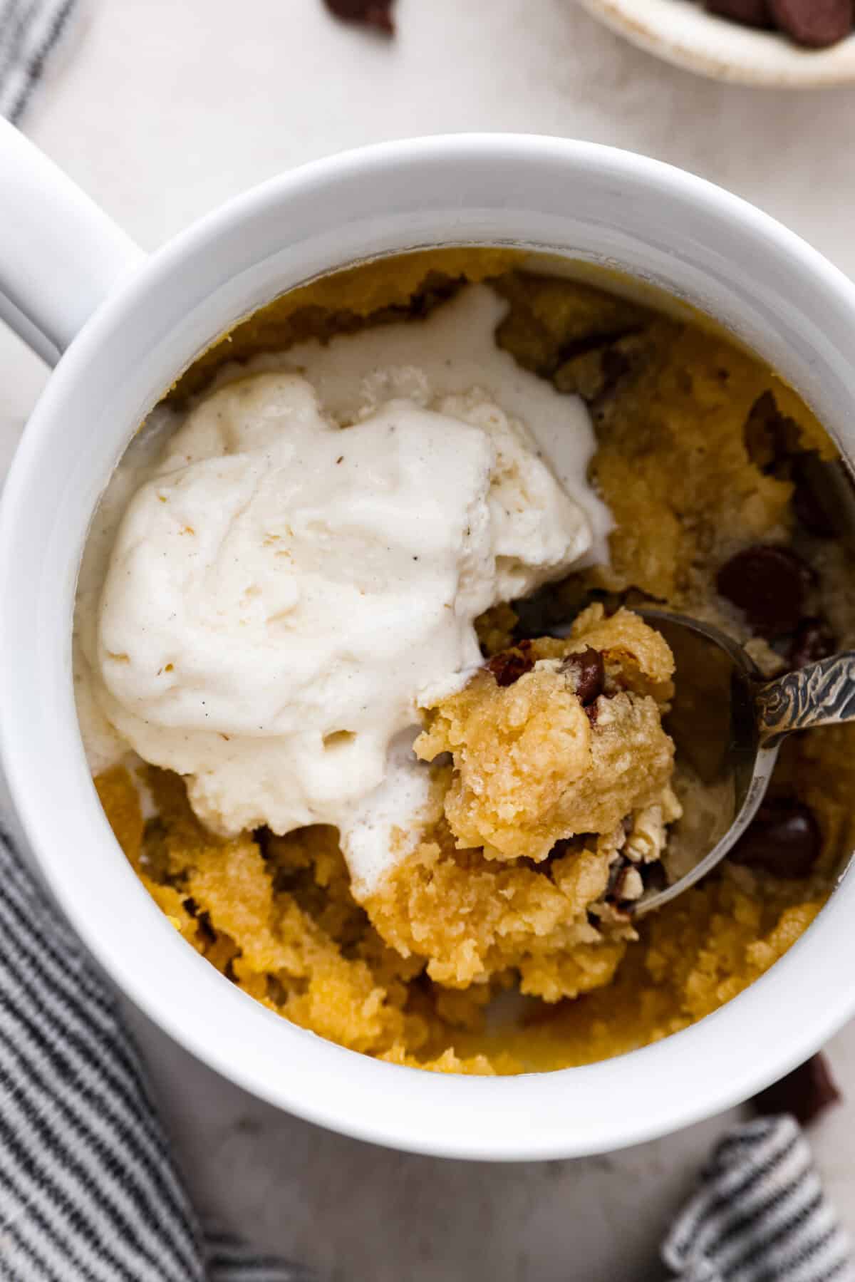 This cookie in a mug recipe is the perfect way to satisfy your cookie craving. In just a few minutes, you can enjoy a warm, buttery, ooey-gooey cookie right in your own mug. Top it with a scoop of ice cream or a dollop of whipped cream, and you'll find yourself in cookie heaven! Close up shot of someone scooping a bite with a bit of ice cream with a spoon.