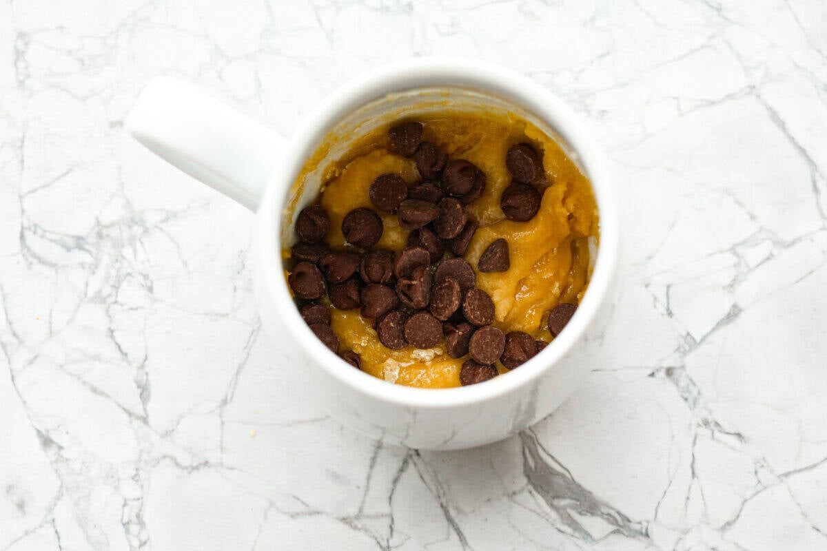 This cookie in a mug recipe is the perfect way to satisfy your cookie craving. In just a few minutes, you can enjoy a warm, buttery, ooey-gooey cookie right in your own mug. Top it with a scoop of ice cream or a dollop of whipped cream, and you'll find yourself in cookie heaven! Overhead shot of mug with cookie dough and chocolate chips dded.