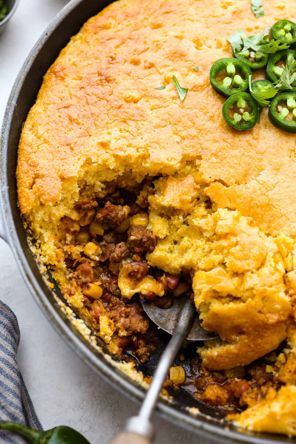 Cowboy Cornbread Casserole is a flavorful, comforting meal that's so easy to make. Seasoned ground beef, corn, pinto beans, and Rotel tomatoes are layered with shredded cheddar cheese and a cornbread topping for a winning dinner! Close up shot of cowboy cornbread casserole dish with spoon inside.