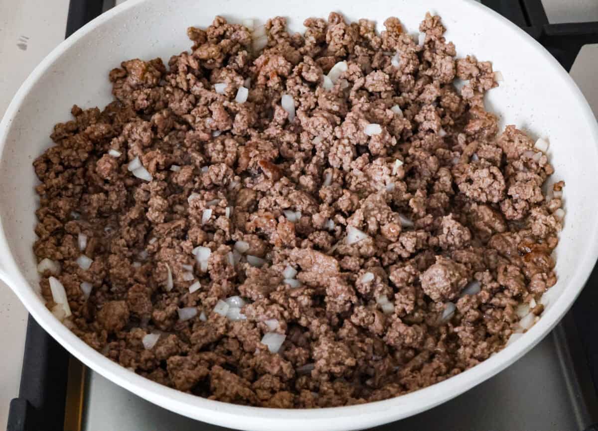 Cowboy Cornbread Casserole is a flavorful, comforting meal that's so easy to make. Seasoned ground beef, corn, pinto beans, and Rotel tomatoes are layered with shredded cheddar cheese and a cornbread topping for a winning dinner! Angle shot of ground beef with onions cooking on the stove top in a skillet.