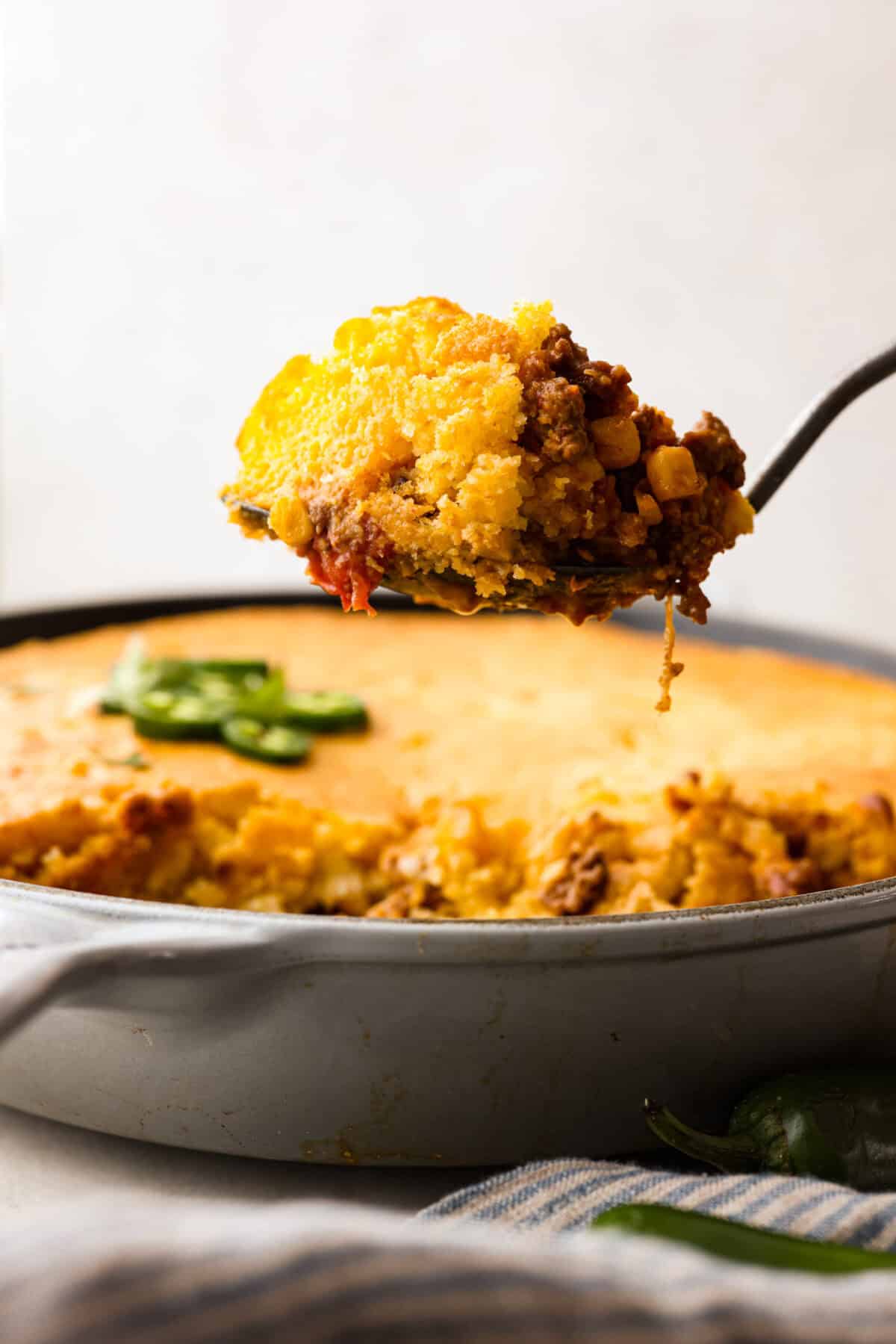 Cowboy Cornbread Casserole is a flavorful, comforting meal that's so easy to make. Seasoned ground beef, corn, pinto beans, and Rotel tomatoes are layered with shredded cheddar cheese and a cornbread topping for a winning dinner! Side shot of a spoon holding a scoop of cowboy cornbread casserole above the whole dish.