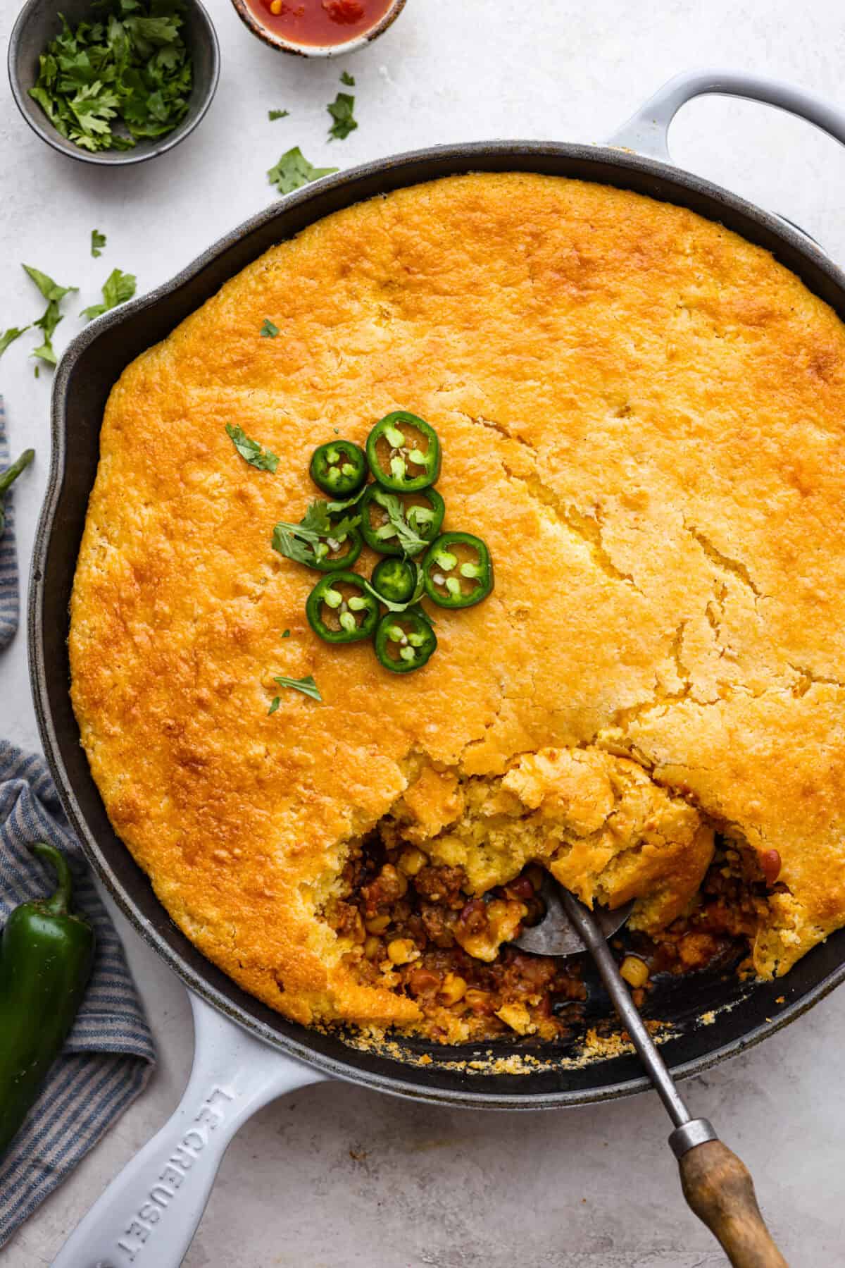Cowboy Cornbread Casserole is a flavorful, comforting meal that's so easy to make. Seasoned ground beef, corn, pinto beans, and Rotel tomatoes are layered with shredded cheddar cheese and a cornbread topping for a winning dinner! Overhead shot of cowboy cornbread casserole with a garnish jalapeños on top and a spoon scooping some out.