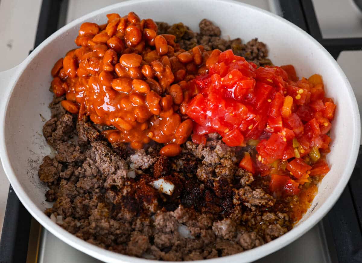 Cowboy Cornbread Casserole is a flavorful, comforting meal that's so easy to make. Seasoned ground beef, corn, pinto beans, and Rotel tomatoes are layered with shredded cheddar cheese and a cornbread topping for a winning dinner! Angle shot of ground beef mixture with can of beans and Rotel dumped over the top.