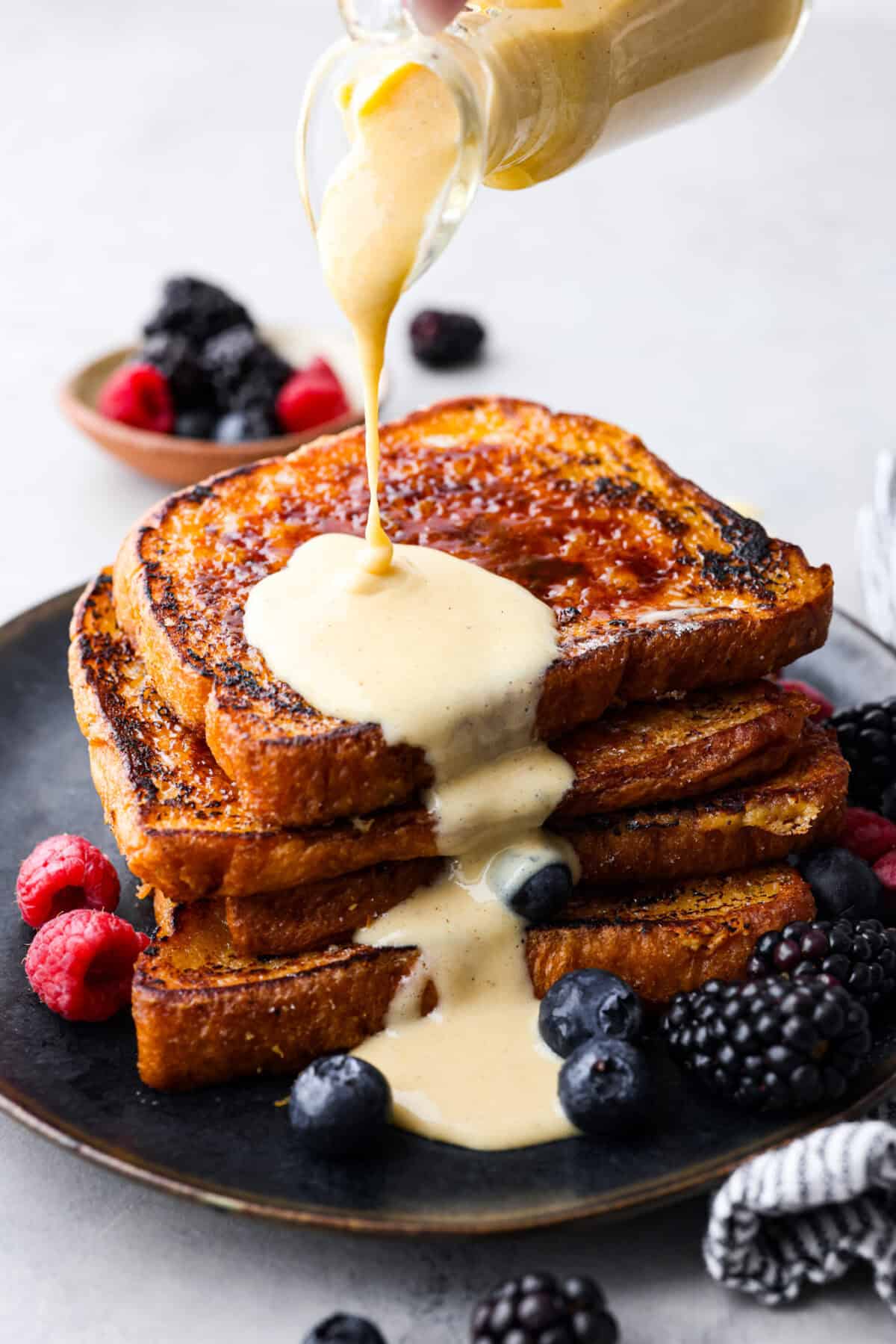 Decadent crème brûlée French toast is fluffy slices of brioche bread with a sweet golden crème brûlée topping. The creme anglaise sauce elevates the French toast making this an unforgettable breakfast! Close view of the creme anglaise sauce pouring on a pile of creme brûlée French toast.