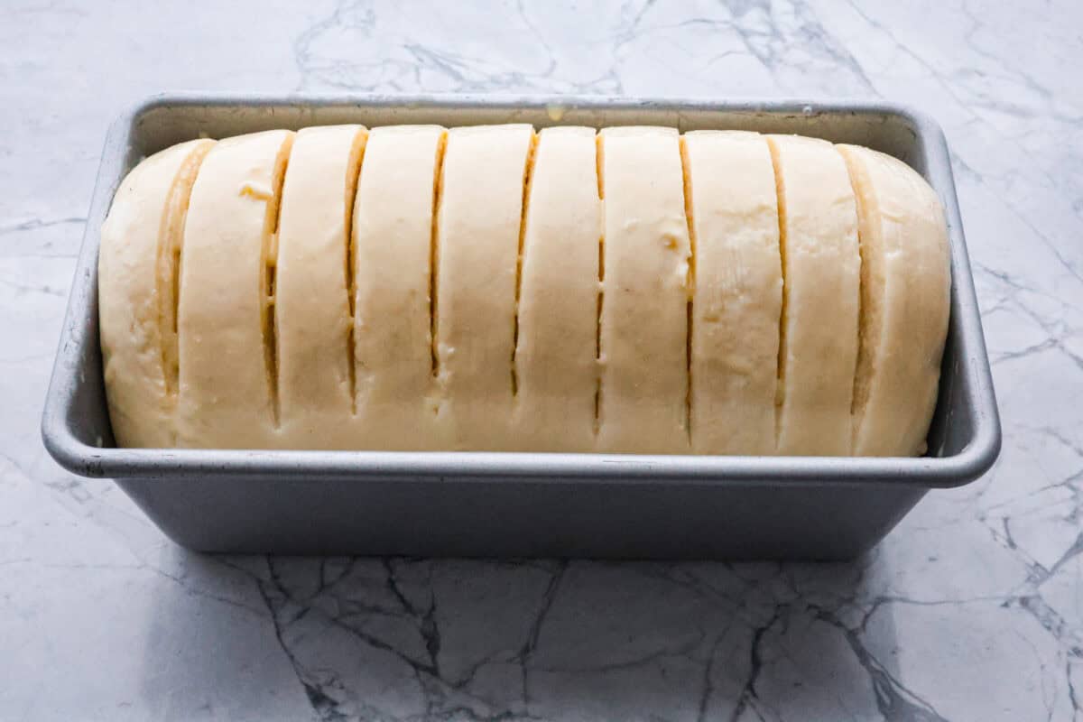 Croissant Bread is beautiful and fluffy with crispy, flaky layers on the outside. It's also one of the most delicious breads you'll ever taste - it will be love at first bite! Add it to your next brunch spread and use it to make French toast, sandwiches, and breakfast casseroles! Overhead shot of scored rolled dough in loaf pan.