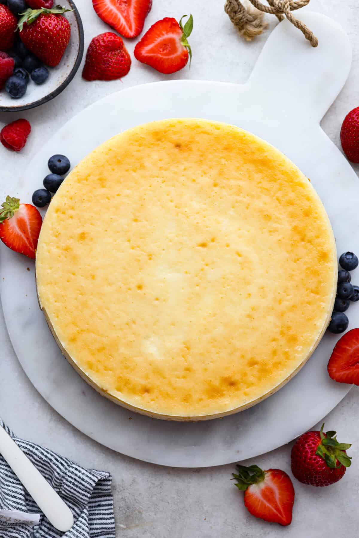 Cheesecake is my absolute FAVORITE dessert! I have perfected my method and I can't wait to teach you how to make cheesecake in this ultimate step-by-step guide. You're on your way to being a cheesecake-making pro! Top view of a whole baked cheesecake with berries on the side.
