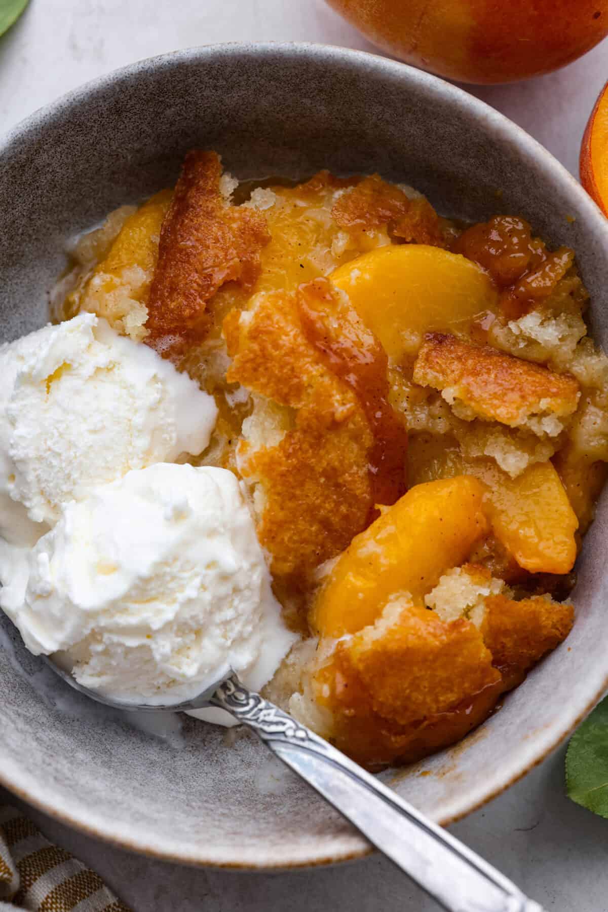 Old-fashioned peach cobbler is a timeless summer dessert! Its warm, cakey layers baked with freshly sliced peaches are a match made in heaven. Top the cobbler with vanilla ice cream, and every bite will be a delight! Overhead shot of a bowl of old fashioned peach cobbler with a scoop of vanilla ice cream.