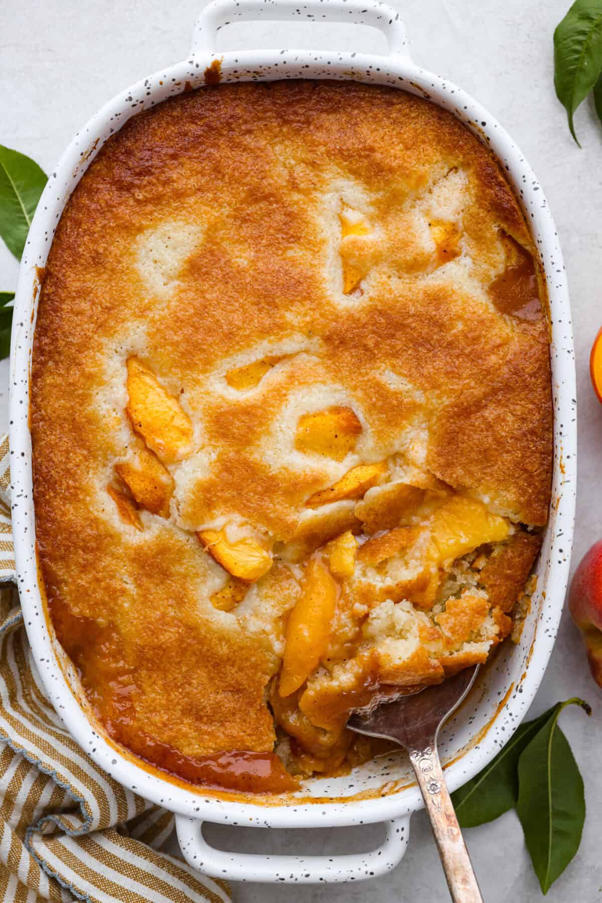Old-fashioned peach cobbler is a timeless summer dessert! Its warm, cakey layers baked with freshly sliced peaches are a match made in heaven. Top the cobbler with vanilla ice cream, and every bite will be a delight! Overhead shot of old fashioned peach cobbler in baking dish with serving spoon sticking out.