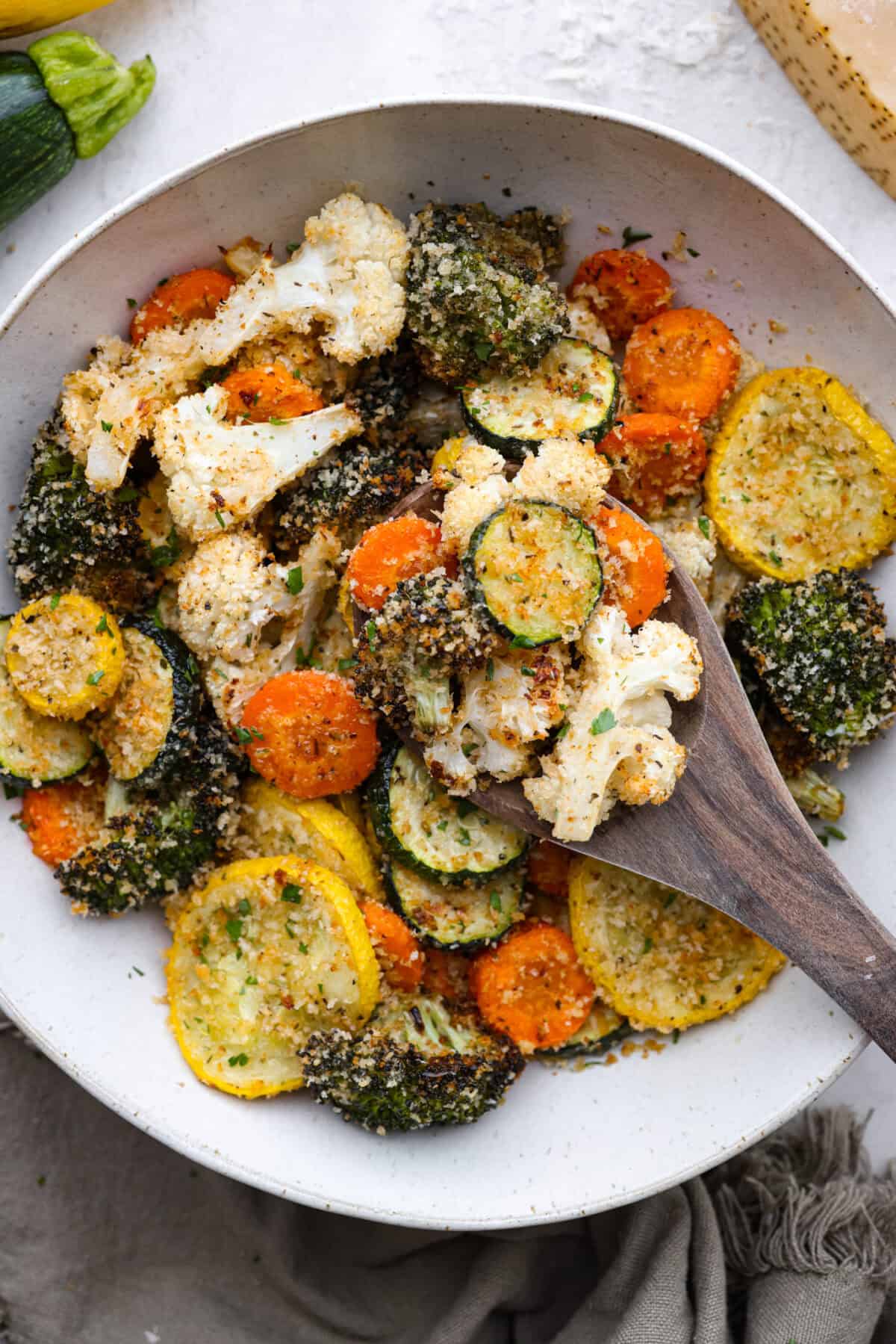 These delicious parmesan roasted vegetables are coated in zesty Italian dressing and a crispy parmesan mixture that delivers incredible flavor. This recipe cooks quickly and works with any vegetable you have on hand, whether using zucchini, cauliflower, or squash. Simply choose your favorite veggies! Overhead shot of parmesan roasted vegetables with wooden spoon.