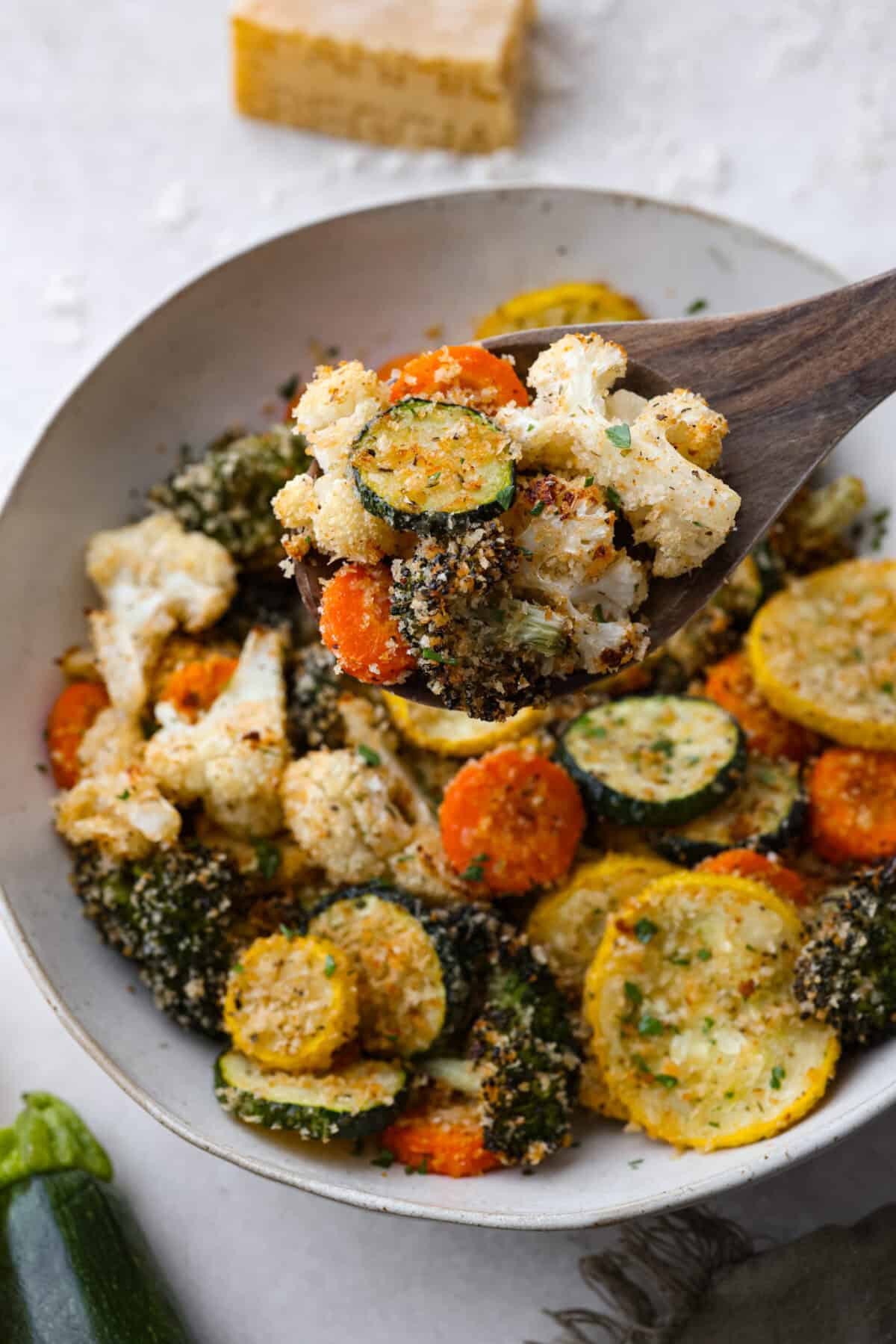 These delicious parmesan roasted vegetables are coated in zesty Italian dressing and a crispy parmesan mixture that delivers incredible flavor. This recipe cooks quickly and works with any vegetable you have on hand, whether using zucchini, cauliflower, or squash. Simply choose your favorite veggies! Close up shot of a wooden spoon filled with parmesan roasted vegetables.