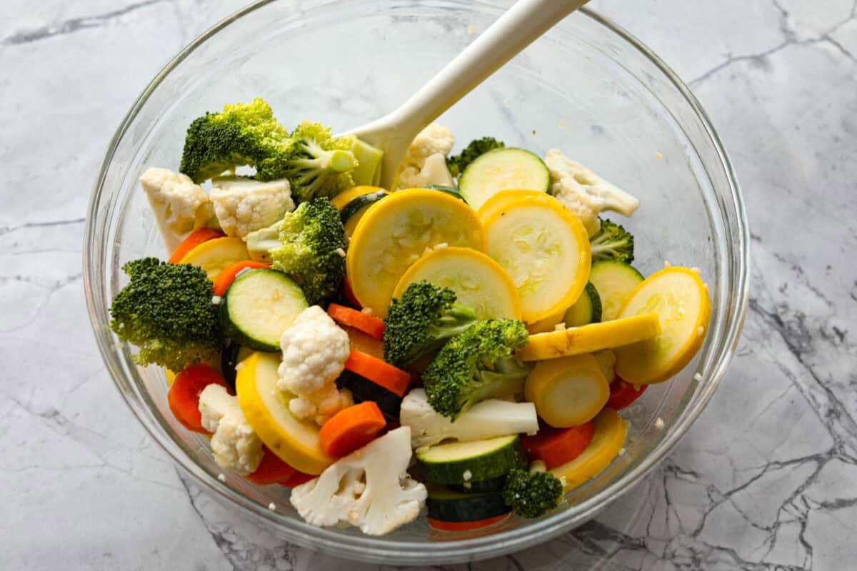 These delicious parmesan roasted vegetables are coated in zesty Italian dressing and a crispy parmesan mixture that delivers incredible flavor. This recipe cooks quickly and works with any vegetable you have on hand, whether using zucchini, cauliflower, or squash. Simply choose your favorite veggies! Overhead shot of vegetables in glass bowl being mixed with olive oil and butter mixture.