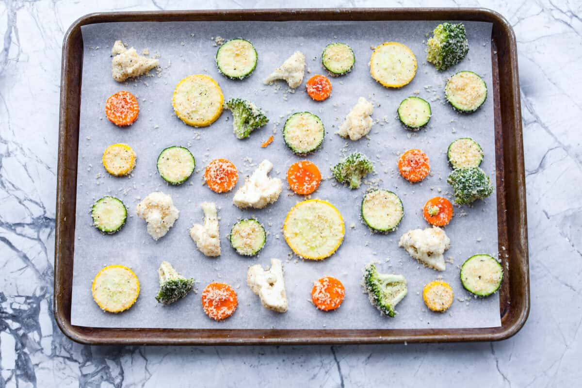 These delicious parmesan roasted vegetables are coated in zesty Italian dressing and a crispy parmesan mixture that delivers incredible flavor. This recipe cooks quickly and works with any vegetable you have on hand, whether using zucchini, cauliflower, or squash. Simply choose your favorite veggies! Overhead shot of breaded vegetables spread out on baking sheet.