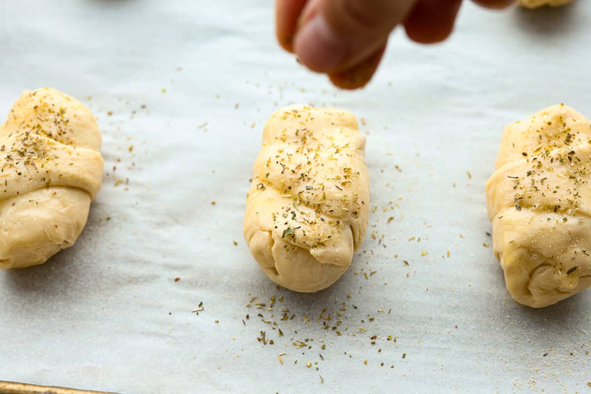 Pizza Stuffed Crescent Rolls areย loaded with gooey mozzarella cheese and pepperoni. Serve with warm marinara for dipping! Angle shot of someone sprinkling Italian seasoning on pizza crescent roll.