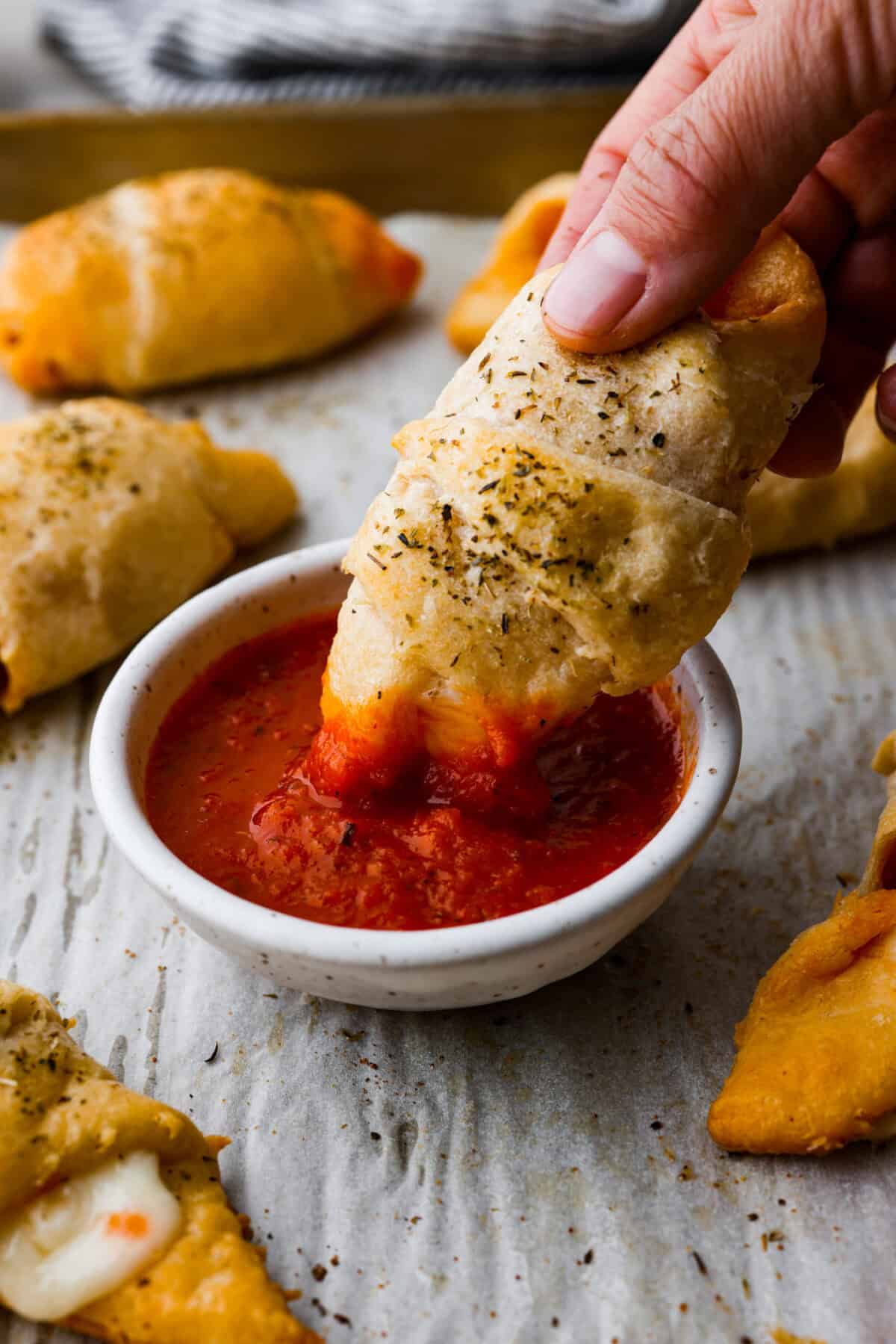 Pizza Stuffed Crescent Rolls areย loaded with gooey mozzarella cheese and pepperoni. Serve with warm marinara for dipping! Angle shot of someone dipping a pizza roll into a small bowl of marinara sauce.