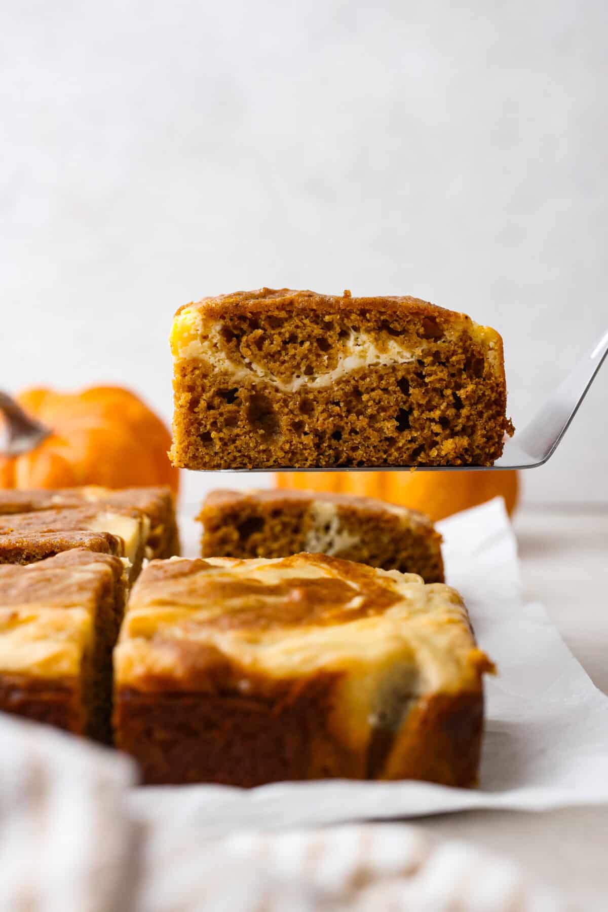 Pumpkin roll bars have all the things you love about pumpkin rolls without all of the hassle! They have perfectly spiced pumpkin cake with a smooth cream cheese swirl that will be sure to make this your new favorite fall dessert! Side shot of a pumpkin roll bar on a spatula being lifted up off a counter, that has parchment paper with other bars on it and small pumpkins in the background.