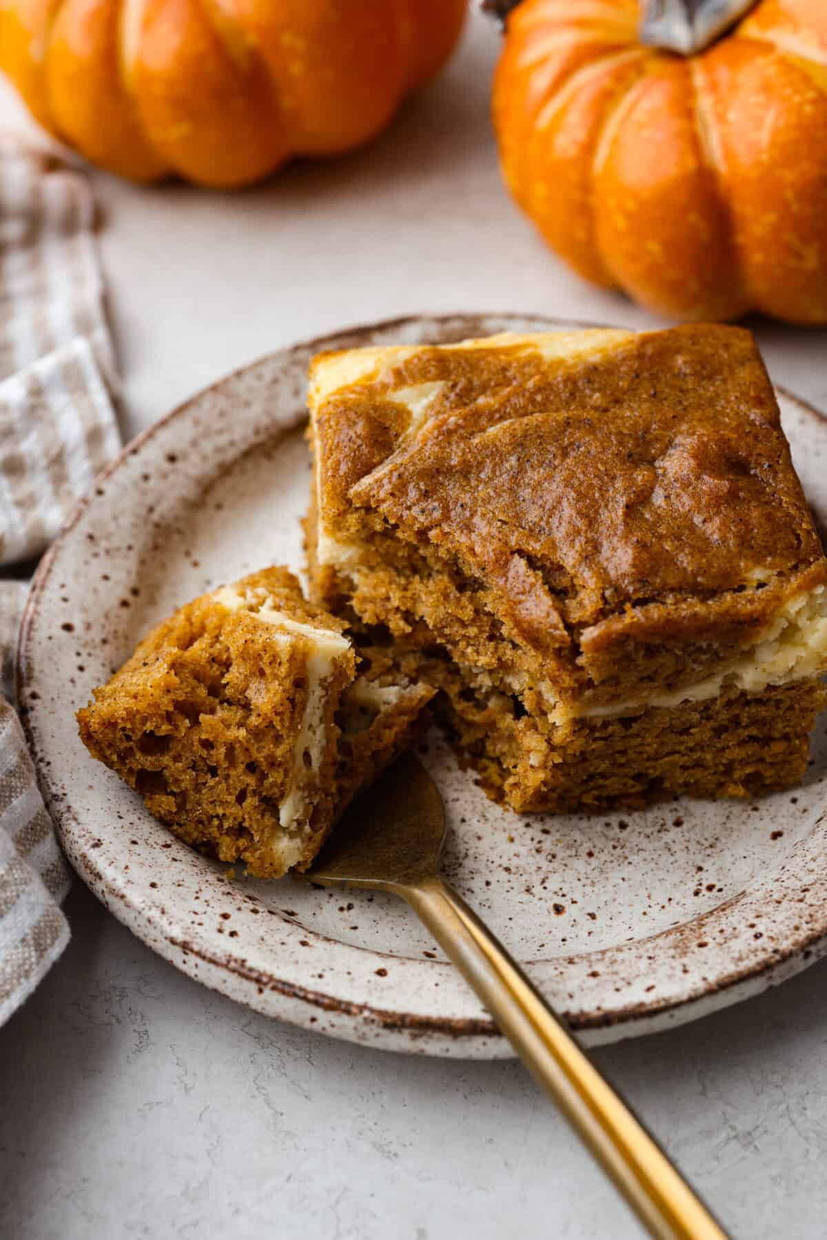 Pumpkin roll bars have all the things you love about pumpkin rolls without all of the hassle! They have perfectly spiced pumpkin cake with a smooth cream cheese swirl that will be sure to make this your new favorite fall dessert! Angle shot of a plate pumpkin roll bar with a bite on a gold spoon.