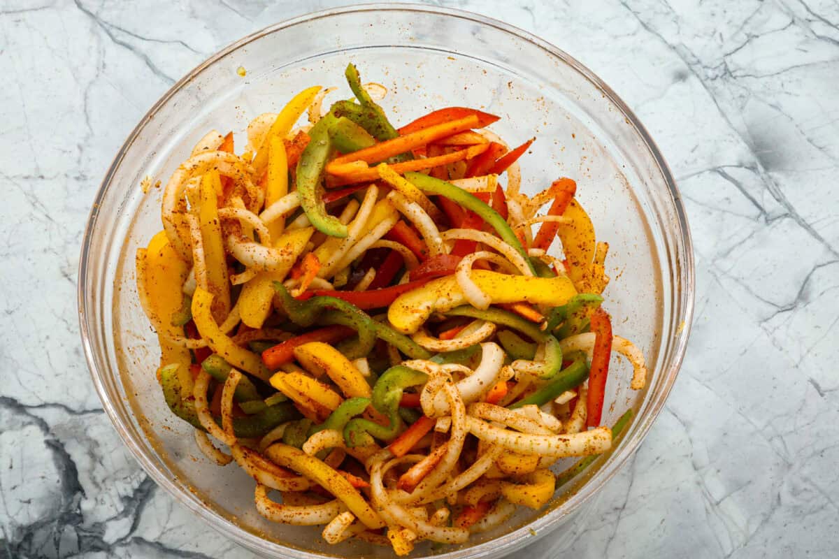 Sheet pan steak fajitas are beautiful and bursting with flavor. Easy to throw together on busy weeknights, they feature tender steak and crisp, colorful vegetables in every bite. You will definitely want to add this to your weekly meal planning! Overhead shot of sliced veggies coated in fajita seasonings in a glass bowl.