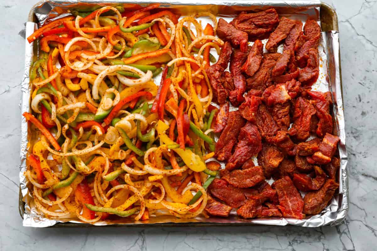 Sheet pan steak fajitas are beautiful and bursting with flavor. Easy to throw together on busy weeknights, they feature tender steak and crisp, colorful vegetables in every bite. You will definitely want to add this to your weekly meal planning! Overhead shot of veggies and steak spread out on a pan.