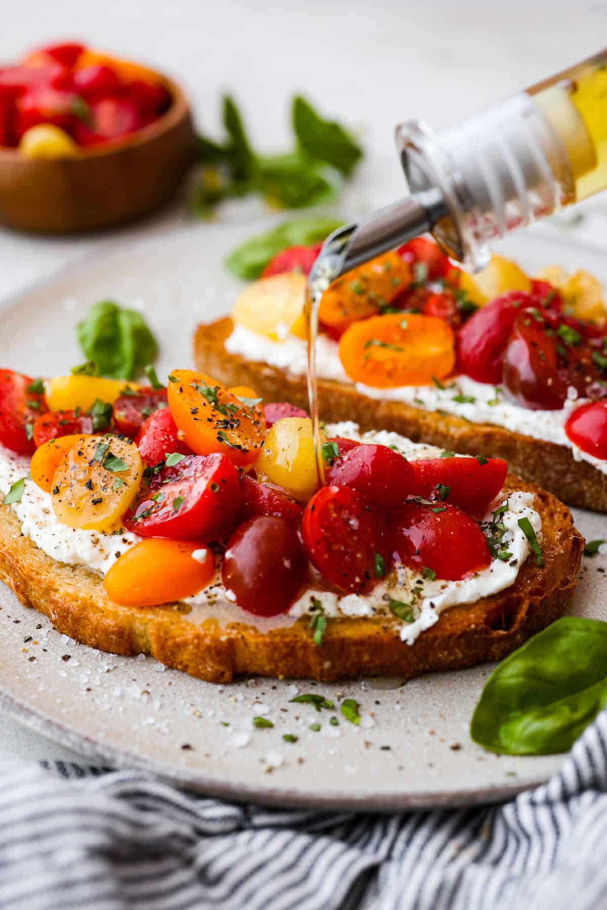 Vibrantย Tomato Toastย has sweet, juicy tomatoes, garlic, creamy ricotta, and fresh basil layered on a crusty slice of toasted bread. Itโs delicious and savory with hints of sweetness. Serve it for breakfast, brunch, as an appetizer, or light lunch for the perfect way to use up summer produce. Side shot of someone drizzling olive oil over tomato toast.