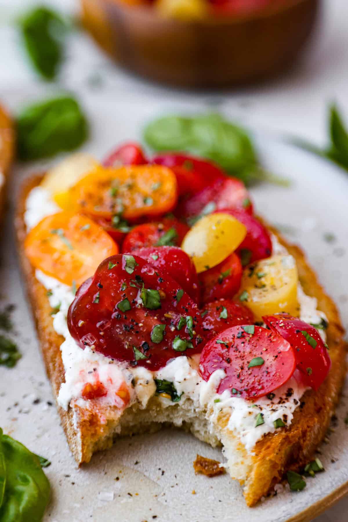 Vibrantย Tomato Toastย has sweet, juicy tomatoes, garlic, creamy ricotta, and fresh basil layered on a crusty slice of toasted bread. Itโs delicious and savory with hints of sweetness. Serve it for breakfast, brunch, as an appetizer, or light lunch for the perfect way to use up summer produce. Angle close up of plated tomato toast with a bite taken out of it.