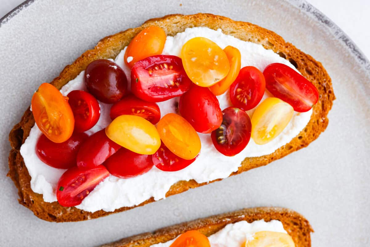 Vibrantย Tomato Toastย has sweet, juicy tomatoes, garlic, creamy ricotta, and fresh basil layered on a crusty slice of toasted bread. Itโs delicious and savory with hints of sweetness. Serve it for breakfast, brunch, as an appetizer, or light lunch for the perfect way to use up summer produce. Overhead shot of toasted bread with ricotta and sliced tomatoes on top.