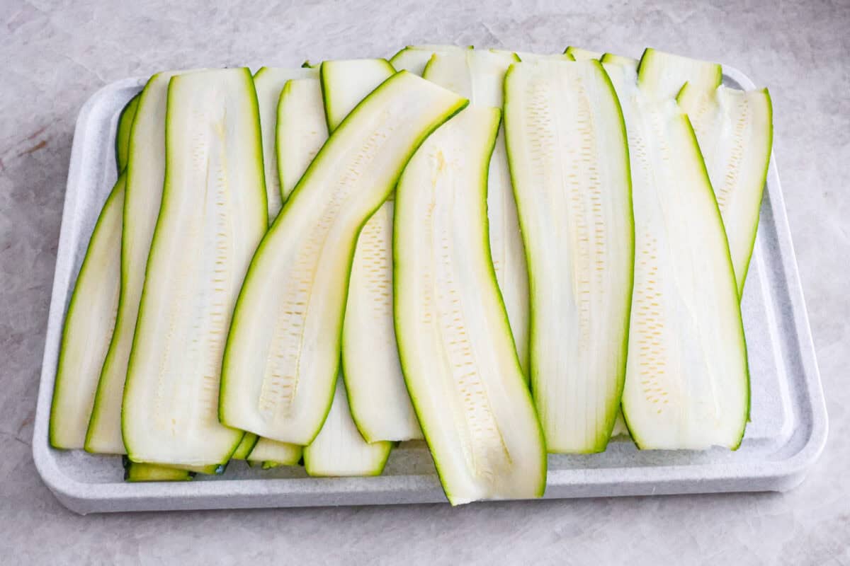 Zucchini Roll-Ups are a delicious, unique meal that the whole family will enjoy. Thin slices of zucchini are rolled around a tasty sausage ricotta mixture and layered over marinara before being topped with cheese and baked to bubbly perfection! Angle shot of zucchini ribbons on cutting board.