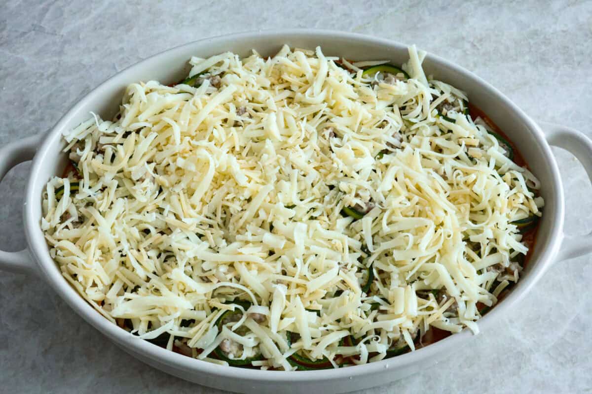 Zucchini Roll-Ups are a delicious, unique meal that the whole family will enjoy. Thin slices of zucchini are rolled around a tasty sausage ricotta mixture and layered over marinara before being topped with cheese and baked to bubbly perfection! Angle shot of pan with all of the zucchini roll up ingredients inside topped with cheese.