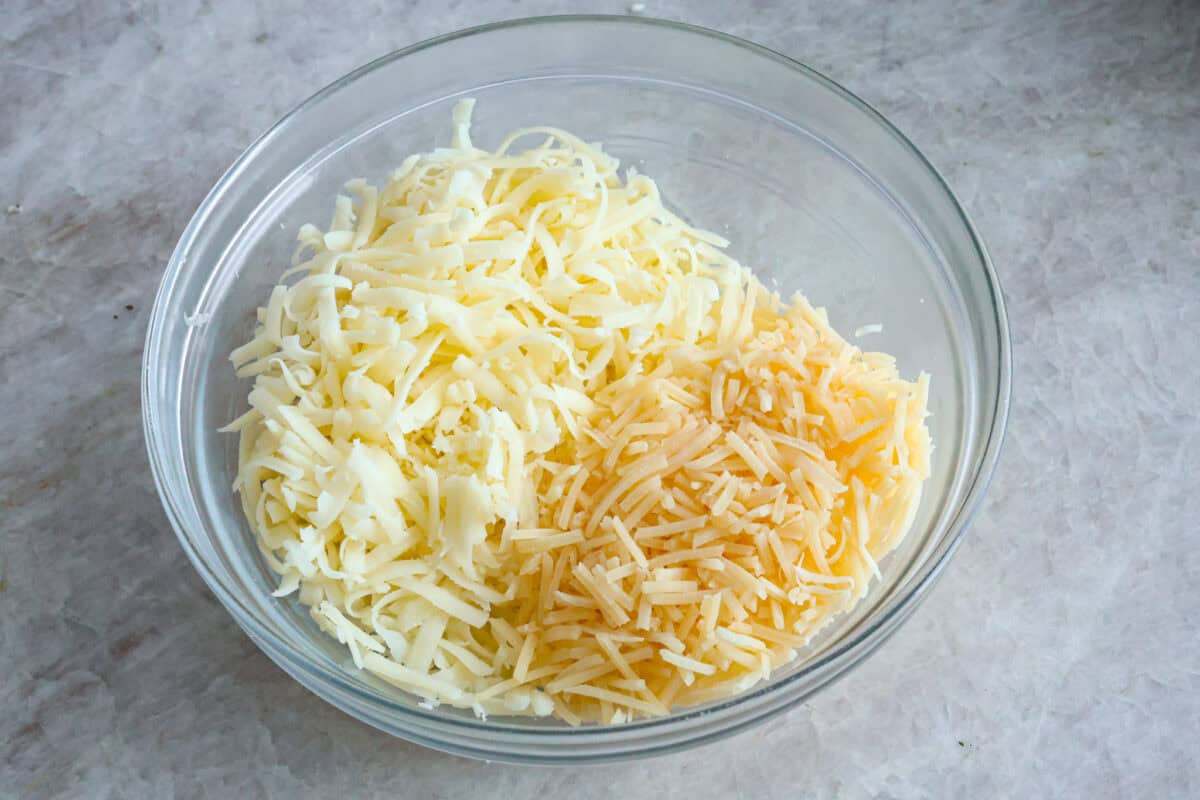Zucchini Roll-Ups are a delicious, unique meal that the whole family will enjoy. Thin slices of zucchini are rolled around a tasty sausage ricotta mixture and layered over marinara before being topped with cheese and baked to bubbly perfection! Angle shot of cheeses in a glass bowl.