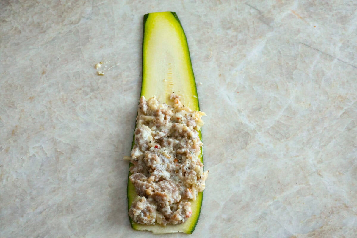 Zucchini Roll-Ups are a delicious, unique meal that the whole family will enjoy. Thin slices of zucchini are rolled around a tasty sausage ricotta mixture and layered over marinara before being topped with cheese and baked to bubbly perfection! Overhead shot of zucchini ribbon with sausage and cheese mixture on one half.