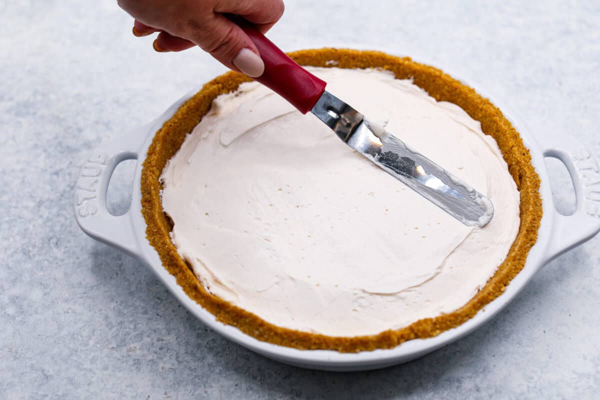 Enjoy juicy, ripe peaches in this Fresh Peach Pie. It has a creamy cheesecake filling topped with sweet peaches, all nestled in a buttery graham cracker crust. Itโs simply delightful, and no baking is required! Angle shot of someone smoothing out the cream filling into the fresh peach pie crust.