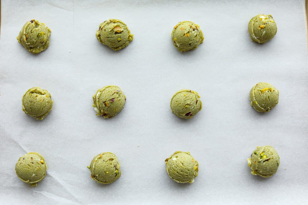 Pistachio Pudding Cookies are super soft, chewy, and irresistible! They're easy to make, and adding pistachio pudding mix gives this cookie recipe wonderful flavor and the perfect tender texture! Overhead shot of cookie dough scooped into balls spread out on a cookie sheet covered in parchment paper.