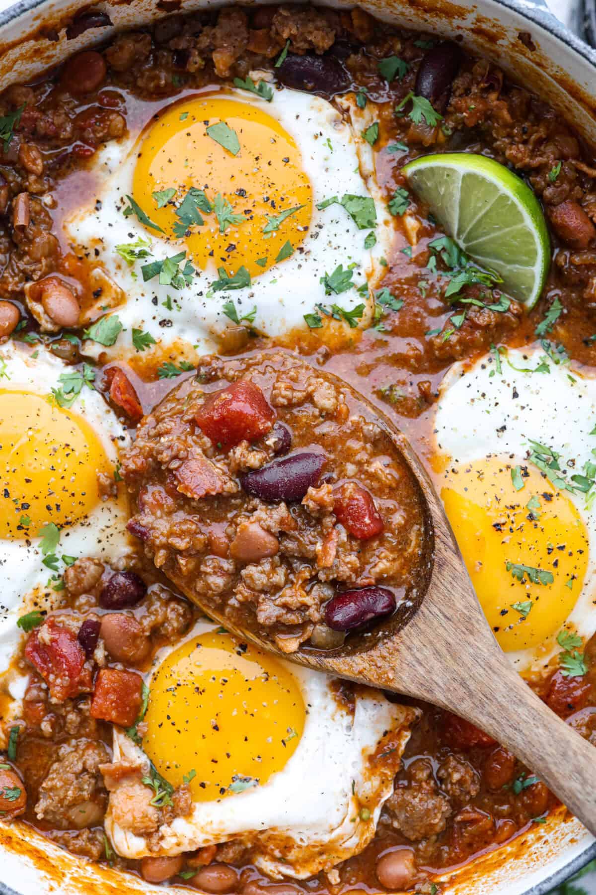 Breakfast Chili is comforting, tasty and will fill you up for hours! A combination of sausage, bacon, tomato sauce, beans, and plenty of tasty seasonings come together for a unique morning meal you will love! Top it off with fried eggs and fresh cilantro for major flavor! Close up shot of someone taking a scoop of breakfast chili out of the pot.