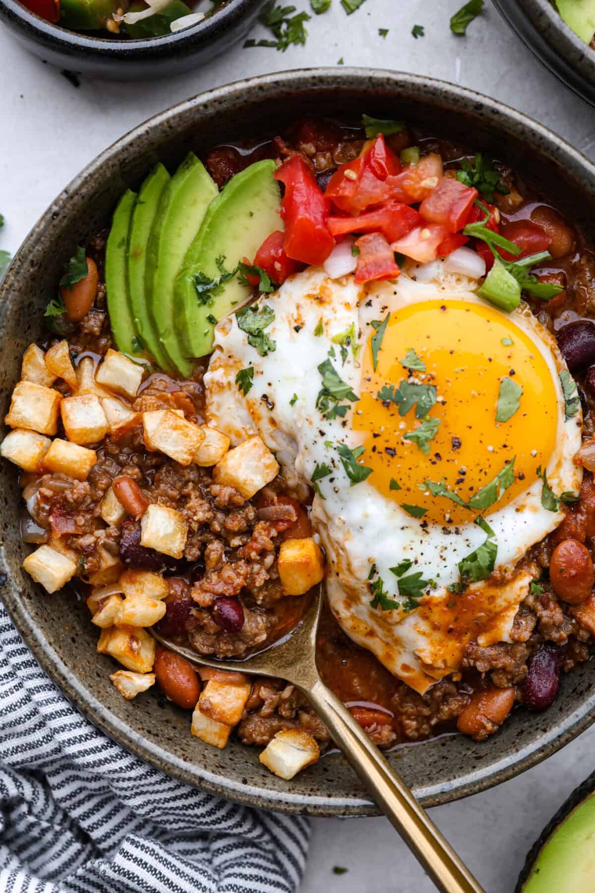 Breakfast Chili is comforting, tasty and will fill you up for hours! A combination of sausage, bacon, tomato sauce, beans, and plenty of tasty seasonings come together for a unique morning meal you will love! Top it off with fried eggs and fresh cilantro for major flavor! Overhead shot of plated breakfast chili with an egg, pico de gallo, cilantro, avocado and hash browns on top.