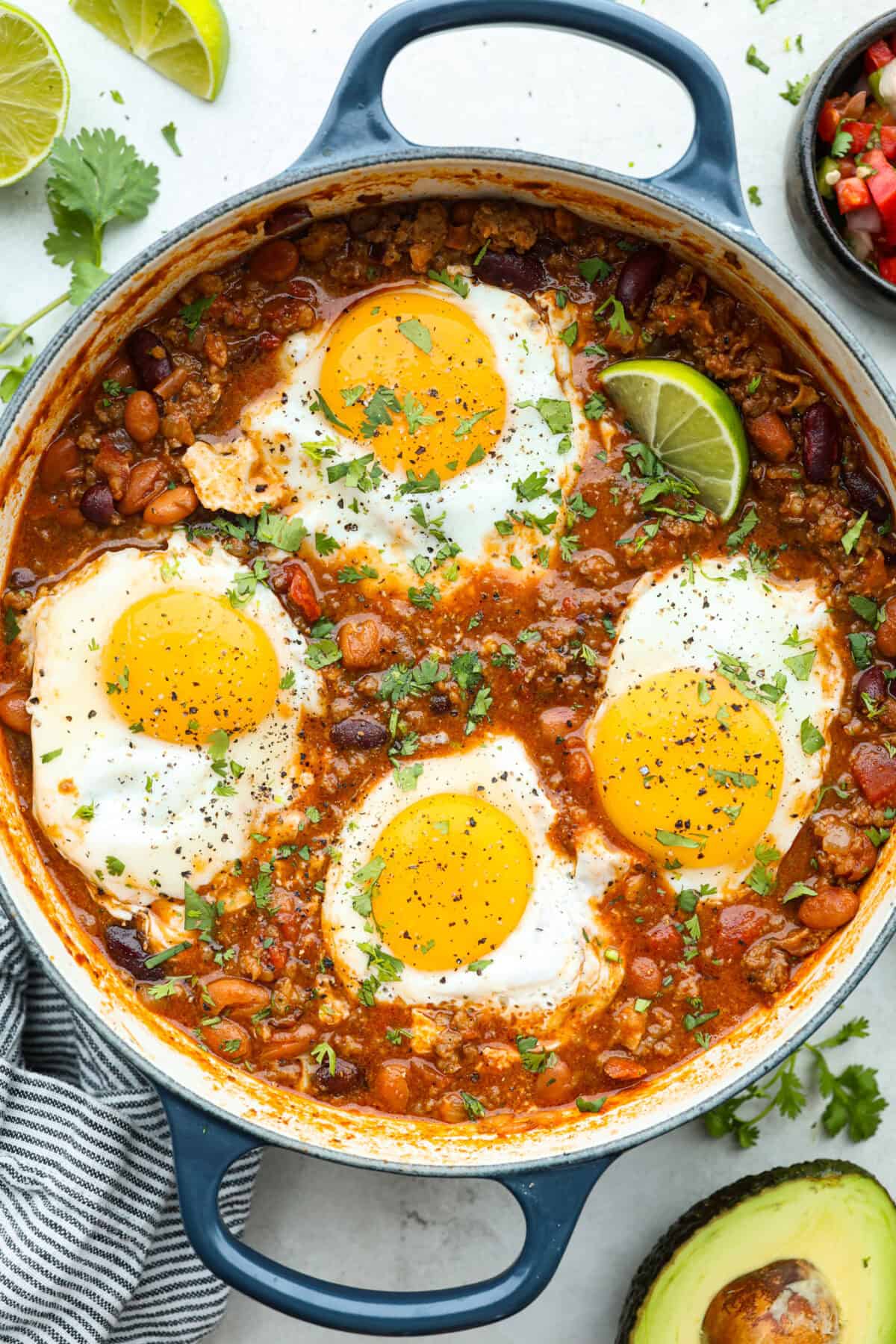 Breakfast Chili is comforting, tasty and will fill you up for hours! A combination of sausage, bacon, tomato sauce, beans, and plenty of tasty seasonings come together for a unique morning meal you will love! Top it off with fried eggs and fresh cilantro for major flavor! Overhead shot of breakfast chili with 4 eggs cooked on top.