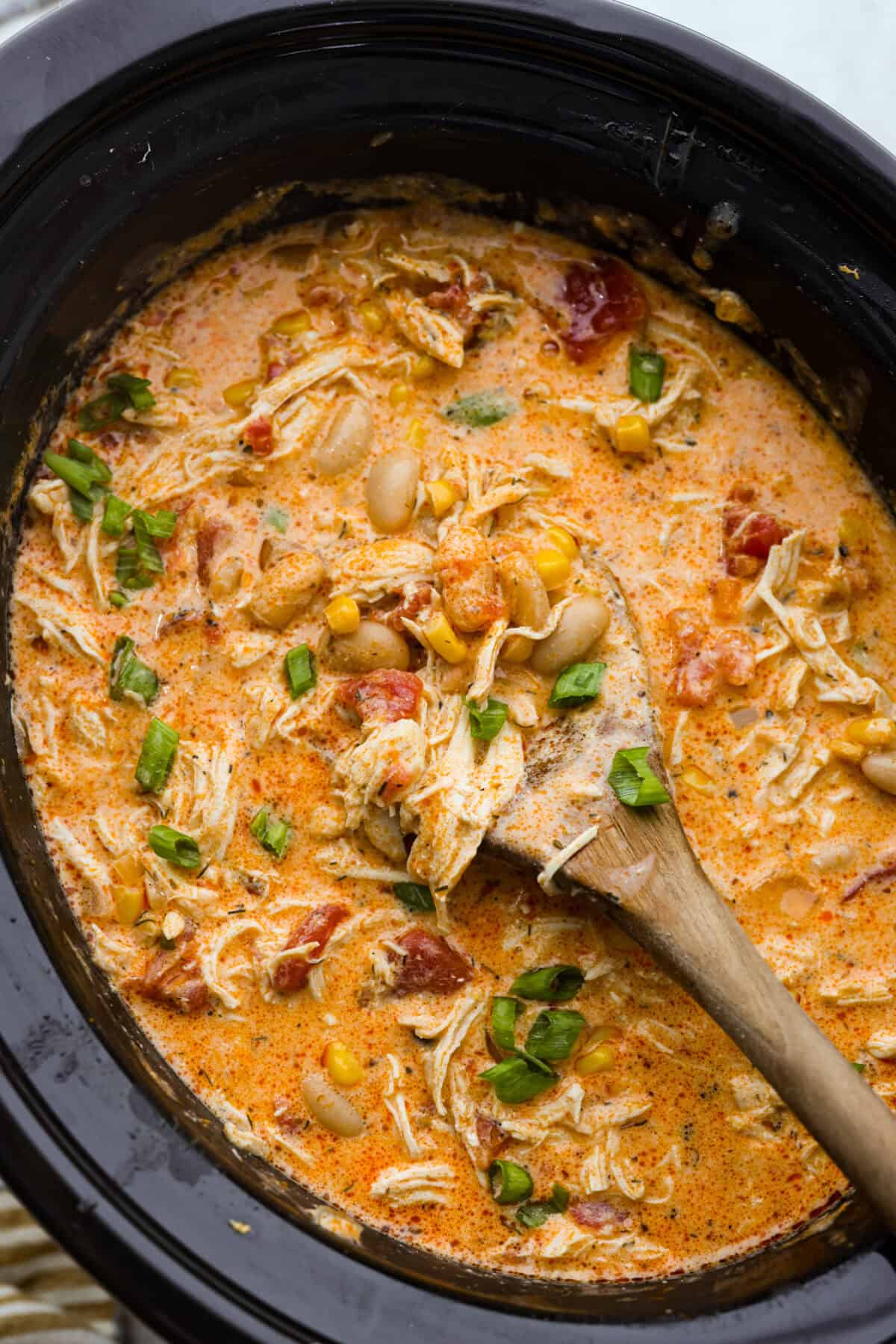 Crockpot buffalo chicken chili is a buffalo wing lover's dream. It has a tangy, fiery buffalo hot sauce base mixed with shredded chicken, smooth cream cheese, tender beans, and vegetables. If you love buffalo wings, you will love this chili! Overhead shot of crockpot buffalo chicken chili with a wooden spoon.