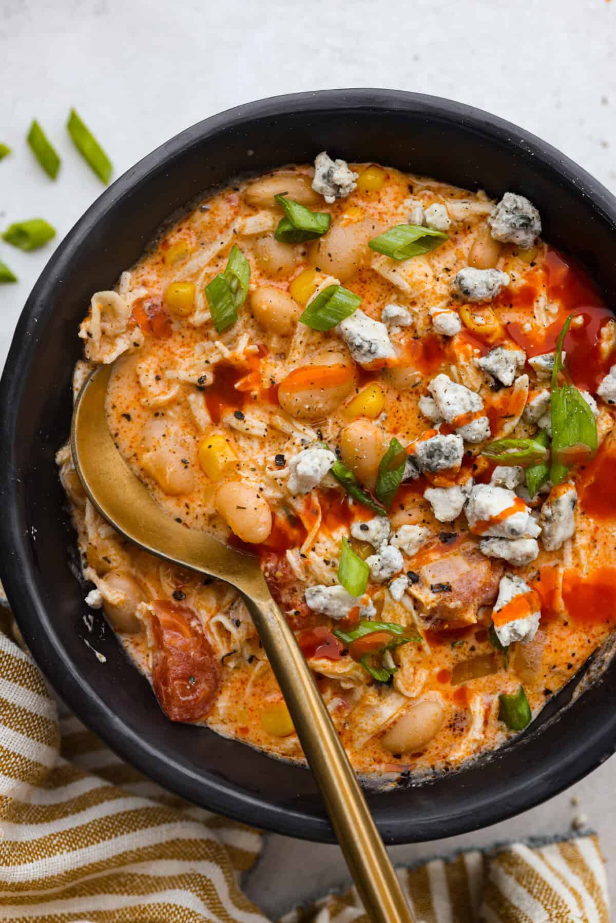 Crockpot buffalo chicken chili is a buffalo wing lover's dream. It has a tangy, fiery buffalo hot sauce base mixed with shredded chicken, smooth cream cheese, tender beans, and vegetables. If you love buffalo wings, you will love this chili! Overhead shot of a bowl of crockpot buffalo chicken chili with a gold spoon, topped with green onion, blue cheese crumbles and a dash more hot suace.
