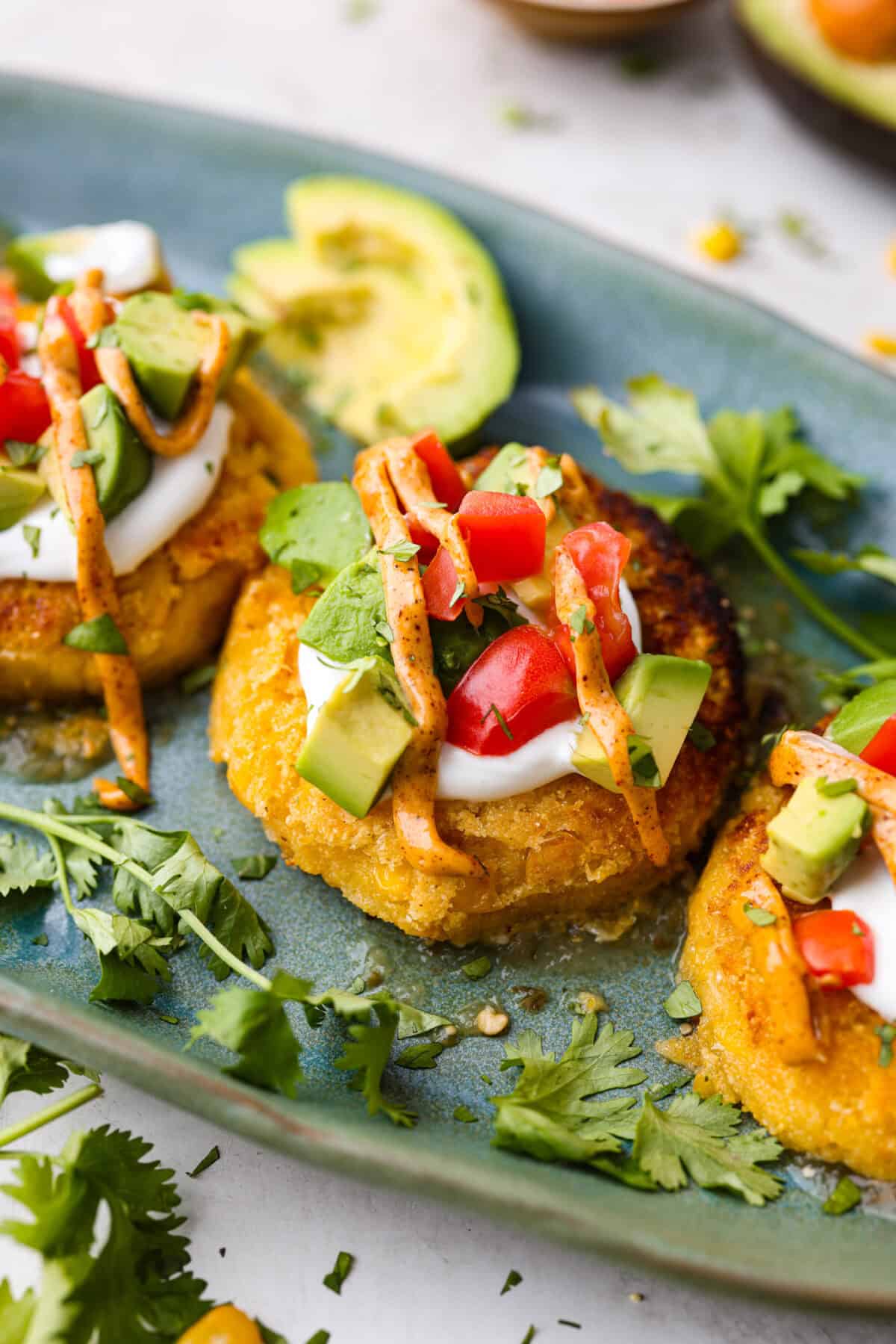Homemade Sweet Corn Tamale Cakes are an explosion of delicious flavor in every bite! With this easy recipe you can bring this Cheesecake Factory favorite right to your own kitchen. Add the signature topping to really take these to the next level! Angle shot of sweet corn tamale cakes plated and topped with fresh tomatoes, avocado, cilantro and sauce.