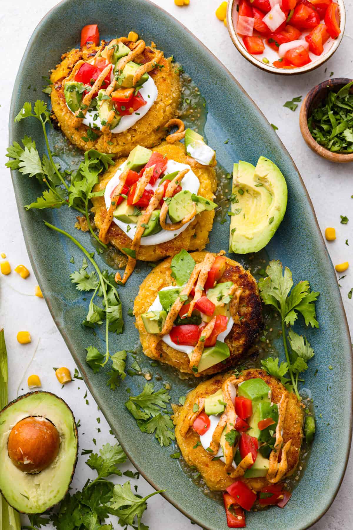 Homemade Sweet Corn Tamale Cakes are an explosion of delicious flavor in every bite! With this easy recipe you can bring this Cheesecake Factory favorite right to your own kitchen. Add the signature topping to really take these to the next level! Overhead shot of 4 sweet corn tamales plated with sauce and toppings and avocado