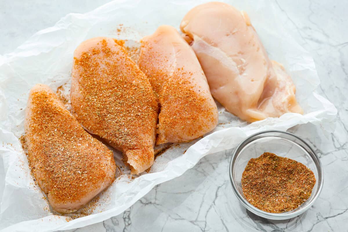 This cowboy chicken recipe is packed with bold flavors. The chicken is seared with lots of vibrant seasonings, surrounded by corn, black beans, and fire-roasted tomatoes, all smothered in melty cheese. Your family will love this quick meal that is ready in just 35 minutes! Overhead shot of chicken breasts being covered in seasonings.