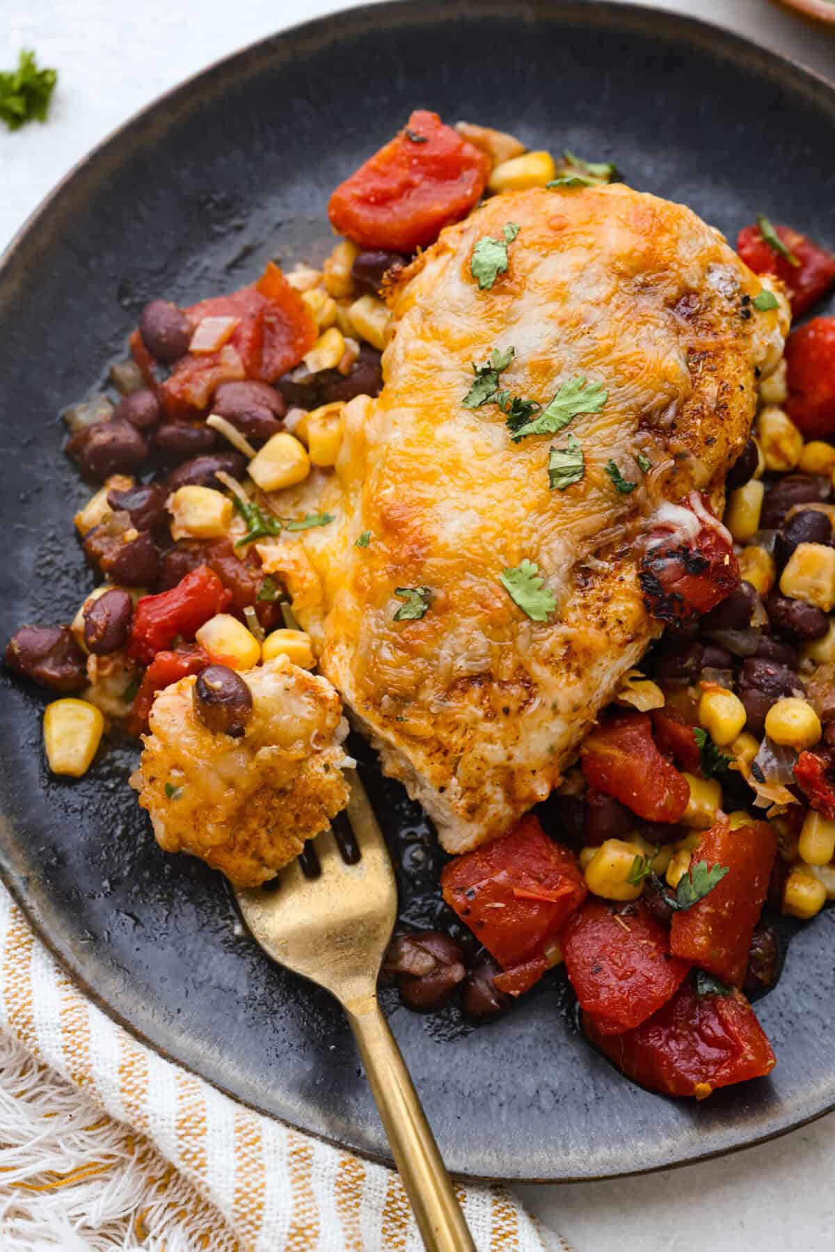 This cowboy chicken recipe is packed with bold flavors. The chicken is seared with lots of vibrant seasonings, surrounded by corn, black beans, and fire-roasted tomatoes, all smothered in melty cheese. Your family will love this quick meal that is ready in just 35 minutes! Overhead shot of plated cowboy chicken with a bit on a gold fork.