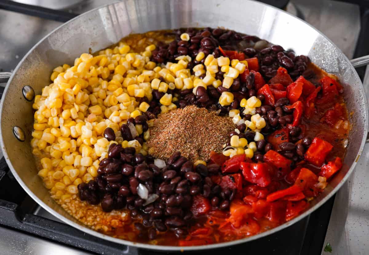 This cowboy chicken recipe is packed with bold flavors. The chicken is seared with lots of vibrant seasonings, surrounded by corn, black beans, and fire-roasted tomatoes, all smothered in melty cheese. Your family will love this quick meal that is ready in just 35 minutes! Angle shot of the corn, beans, tomatoes, onions and seasonings all in a pan.