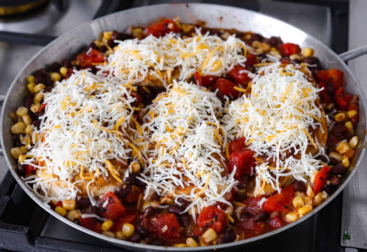 This cowboy chicken recipe is packed with bold flavors. The chicken is seared with lots of vibrant seasonings, surrounded by corn, black beans, and fire-roasted tomatoes, all smothered in melty cheese. Your family will love this quick meal that is ready in just 35 minutes! Close up shot of chicken on top of the tomatoes, beans, corn and onions with sprinkled cheese on top.