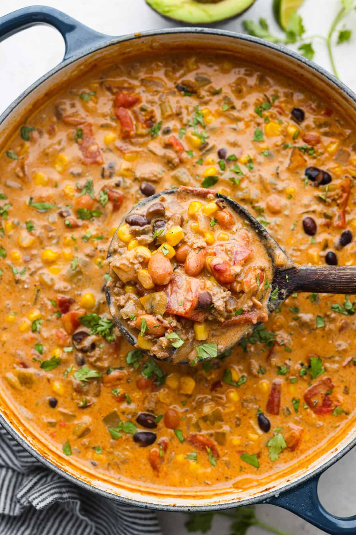 This creamy taco soup recipe is full of fresh flavors, tender vegetables, and hearty beans. The seasoned beef mixed with the creamy soup is out of this world good! Top it with your favorite toppings, and your family will love this warm, savory soup! Overhead shot of a pot of creamy taco soup with a wooden spoon ladling out a scoop.