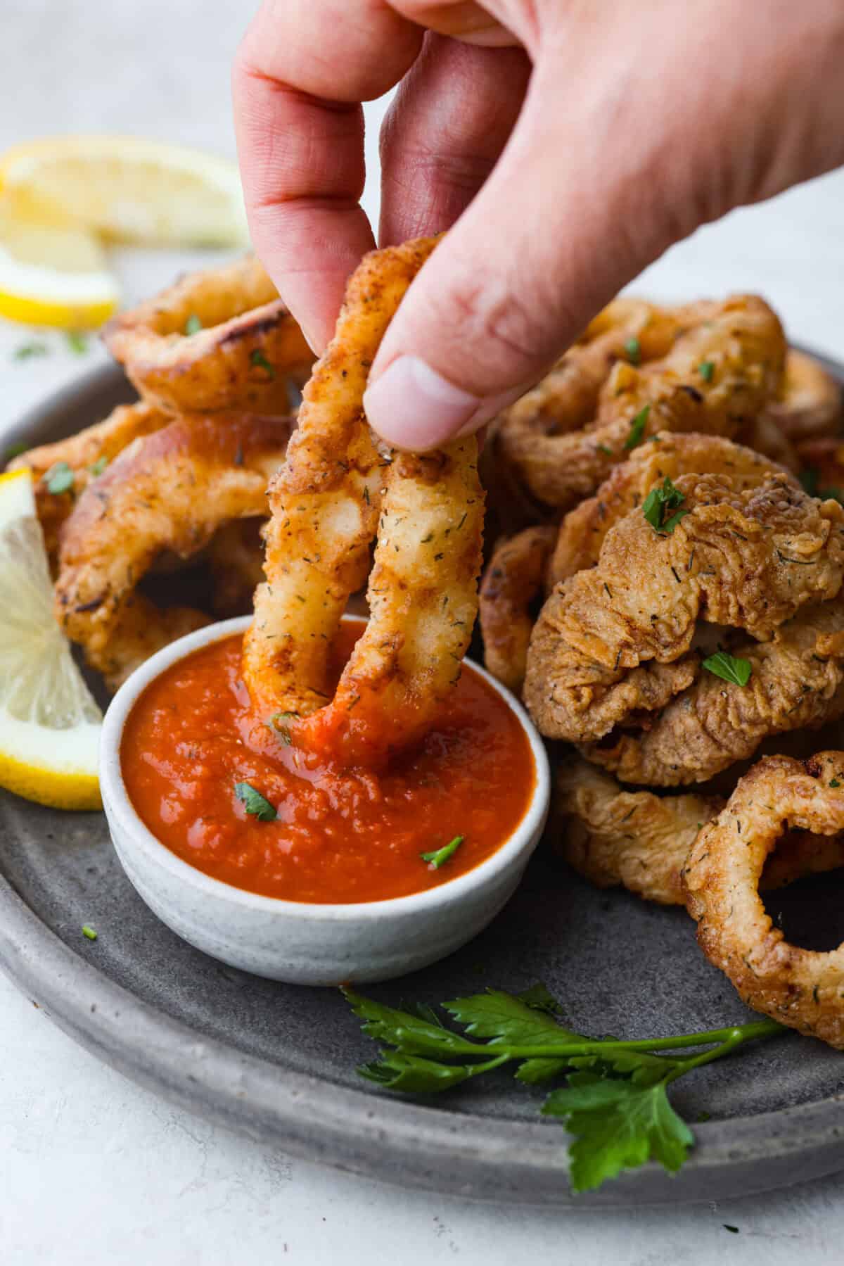 Crispy, flavorful Fried Calamari is a delicious appetizer that tastes like something you would order in a fancy restaurant. The calamari is tenderized, breaded and then fried to golden brown, crunchy perfection. Take it to the next level with marinara sauce for dipping! Side shot of someone dipping a fried calamari ring into marinara sauce.