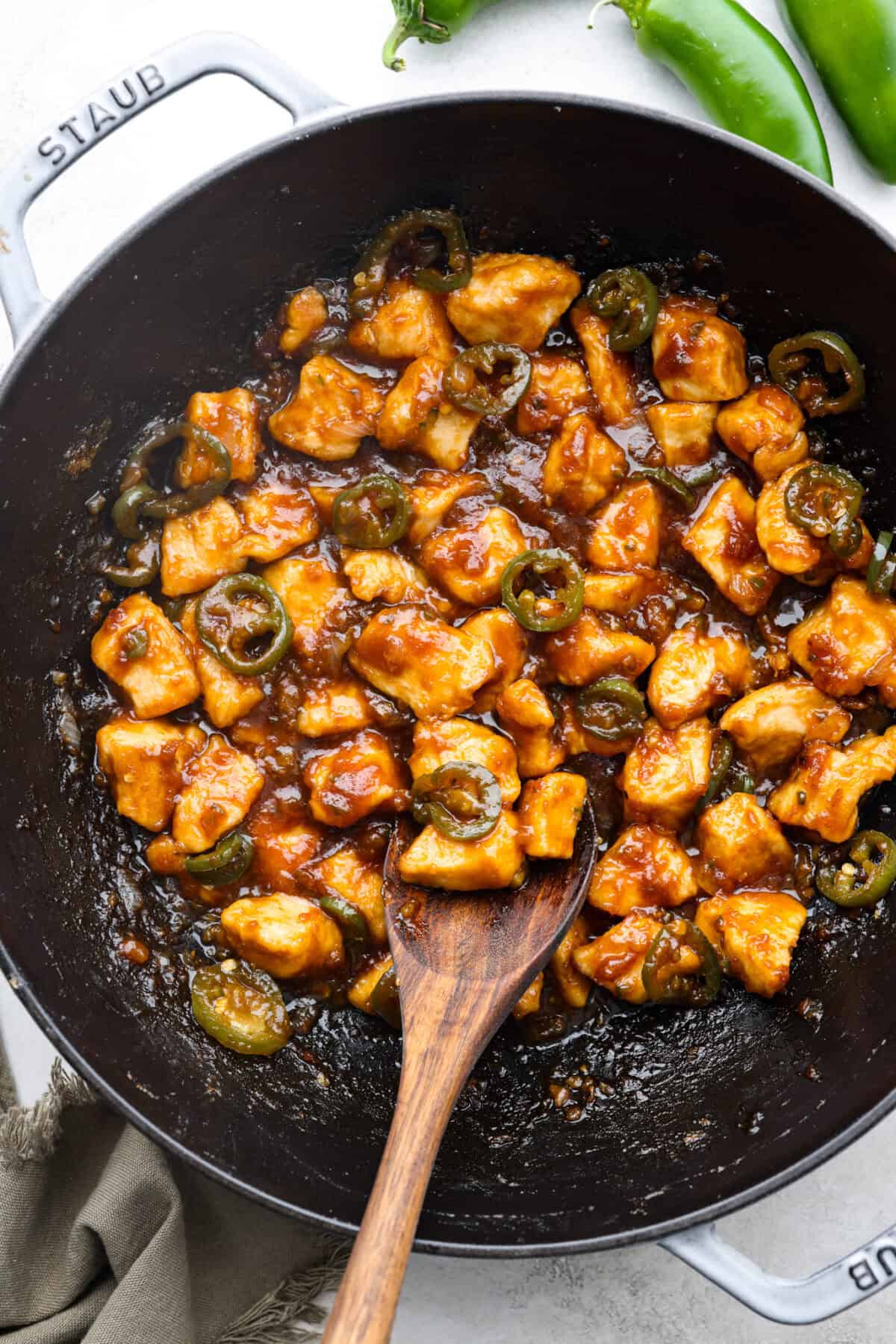 Delicious Jalapeño Chicken will become an instant favorite with the very first bite. Tender chicken pieces are cooked with jalapeños and then coated in a sticky, sweet, and spicy sauce. Serve over rice for an easy meal. You'll be craving it on repeat! Close up shot of jalapeño chicken with a wooden serving spoon in a skillet.