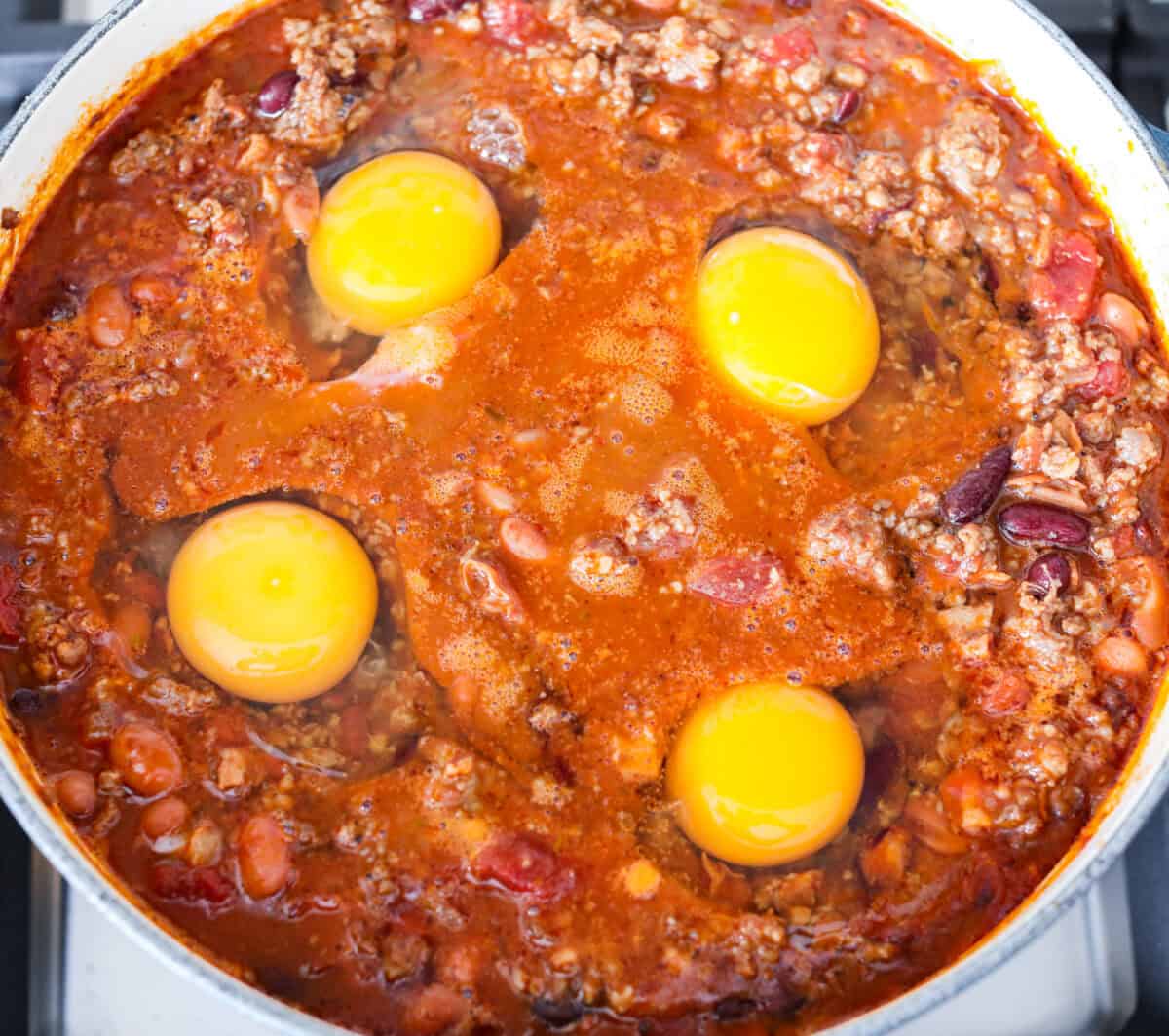 Breakfast Chili is comforting, tasty and will fill you up for hours! A combination of sausage, bacon, tomato sauce, beans, and plenty of tasty seasonings come together for a unique morning meal you will love! Top it off with fried eggs and fresh cilantro for major flavor! Overhead shot of chili with 4 raw eggs over the top.
