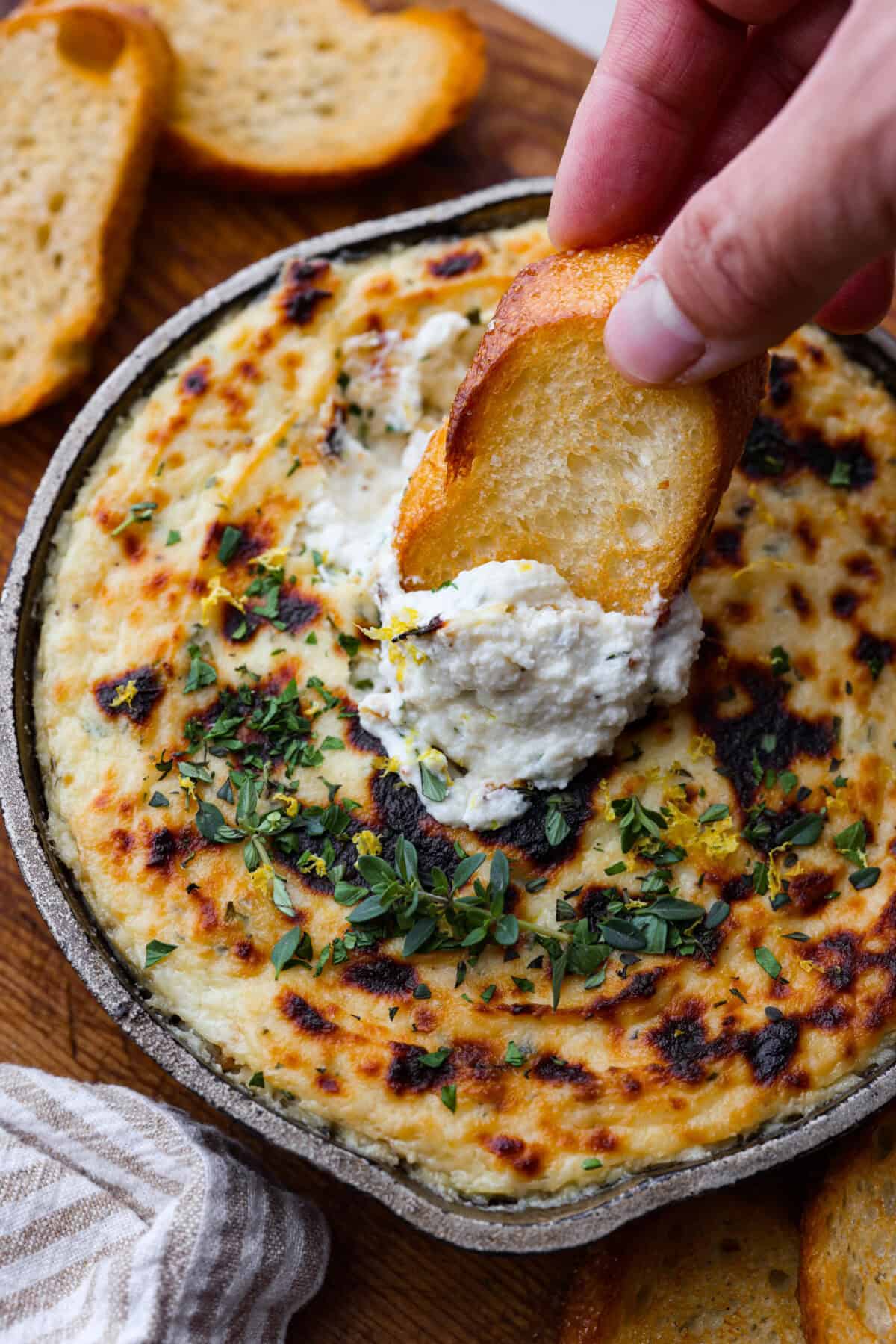 Irresistible Baked Ricotta Dip is the perfect party appetizer! It takes just minutes to combine creamy ricotta cheese with parmesan, fresh herbs, and lemon zest. Then, bake until bubbly and delicious, and get ready for an explosion of flavor! Overhead shot of someone dipping a crostini into the baked ricotta dip.